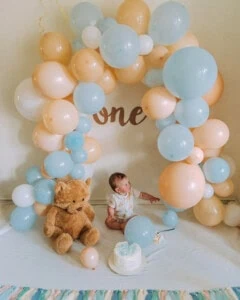 Baby cake smash idea with pastel balloon arch, teddy bear, and layered cake.