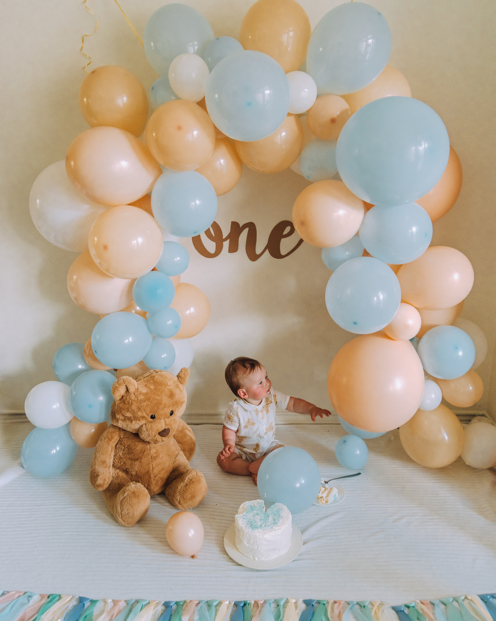 Baby cake smash idea with pastel balloon arch, teddy bear, and layered cake.