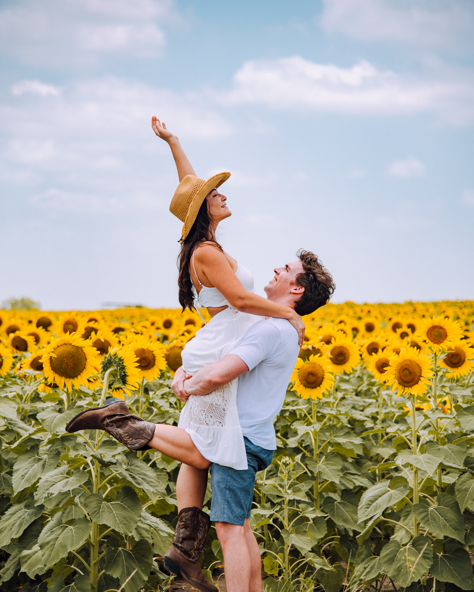 Man holds up a woman wearing a white dress, cowboy hat, and cowboy boots in front of a sunflower field in Texas